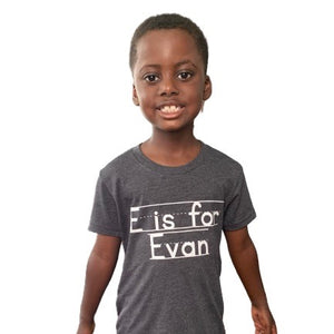 An African American school aged boy smiles while wearing a personalized gray heather T-Shirt that says E is for Evan. Text is written in same font as chalkboard writing or writing practice for elementary students. Shirt is a gift for kids, matching family shirt, holiday kid gift