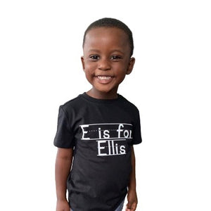 An African American preschool boy smiles while wearing a black personalized T-Shirt that says E is for Ellis.  Text is written in same font as chalkboard writing or writing practice for elementary students. Shirt is a gift for kids, matching family shirt, holiday kid gift