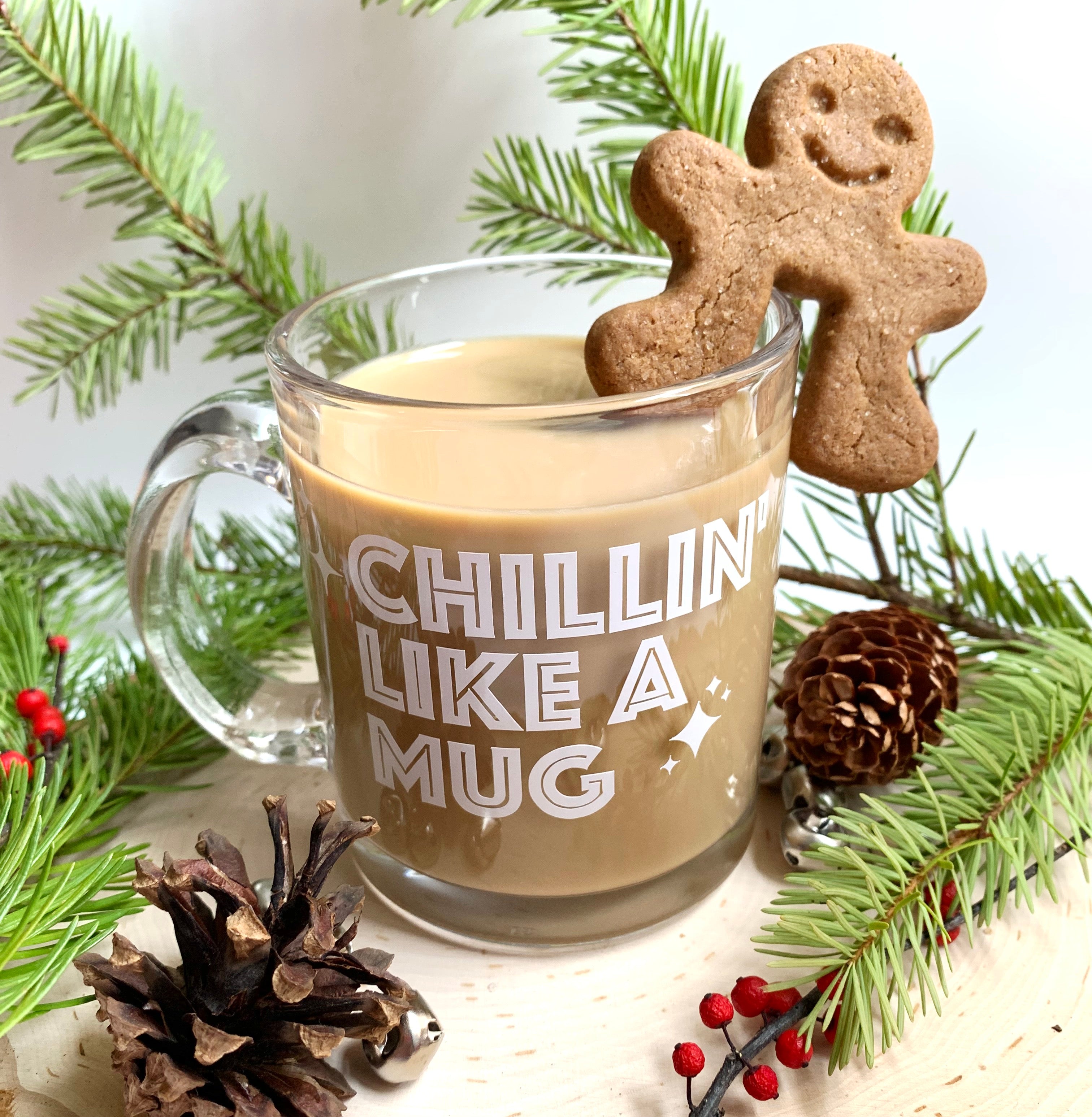 Pictured is clear funny mug that says, Chillin' Like a Mug. The mug site on a round log slice.A gingerbread man is hanging from the mug.  in photo are pinecones, berries and Christmas tree branches. mug is a gift for you, gift for her, gift for him.