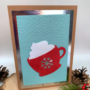 A brown card base with silver backing, embossed falling snow (blue card base), a red mug with a silver snowflake on the front and white whip cream, an sits on the front to the card. the words, "DON WE NOW OUR UGLY SWEATERS" is at the bottom of the card. There is a log slice, jingle bells, pinecone, berries, and Christmas tree branch surround the standing card. Gift for her, gift for him, gift for them