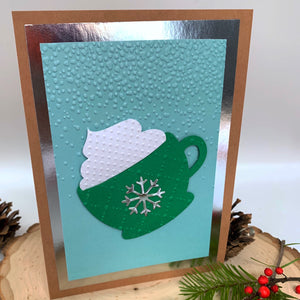 A brown card base with silver backing, embossed falling snow (blue card base), a green mug with a silver snowflake on the front and white whip cream, an sits on the front to the ca. There is a log slice, jingle bells, pinecone, berries, and Christmas tree branch surround the standing card. Gift for her, gift for him, gift for them